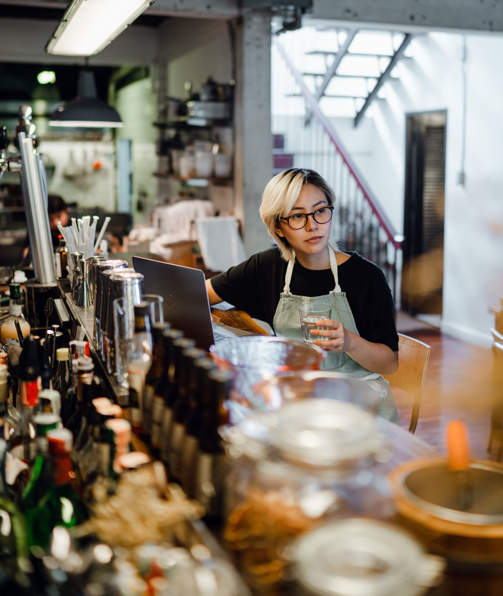 A bar manager in an apron working on a laptop at the end of the bar during a quiet spell.