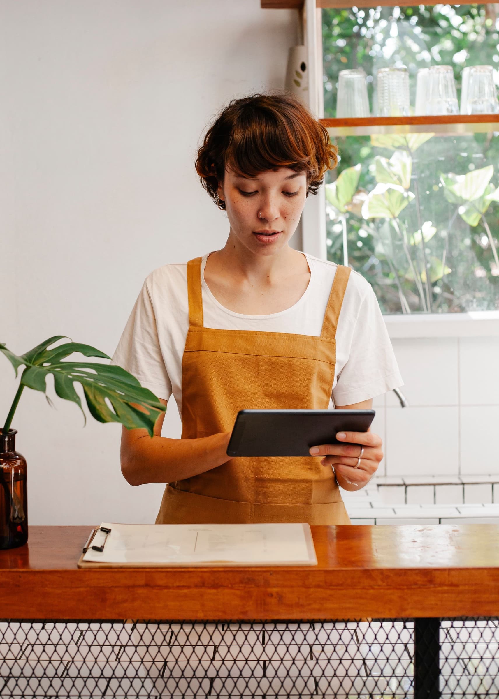 A café worker in an apron checking a tablet at the counter, a clipboard beside her.