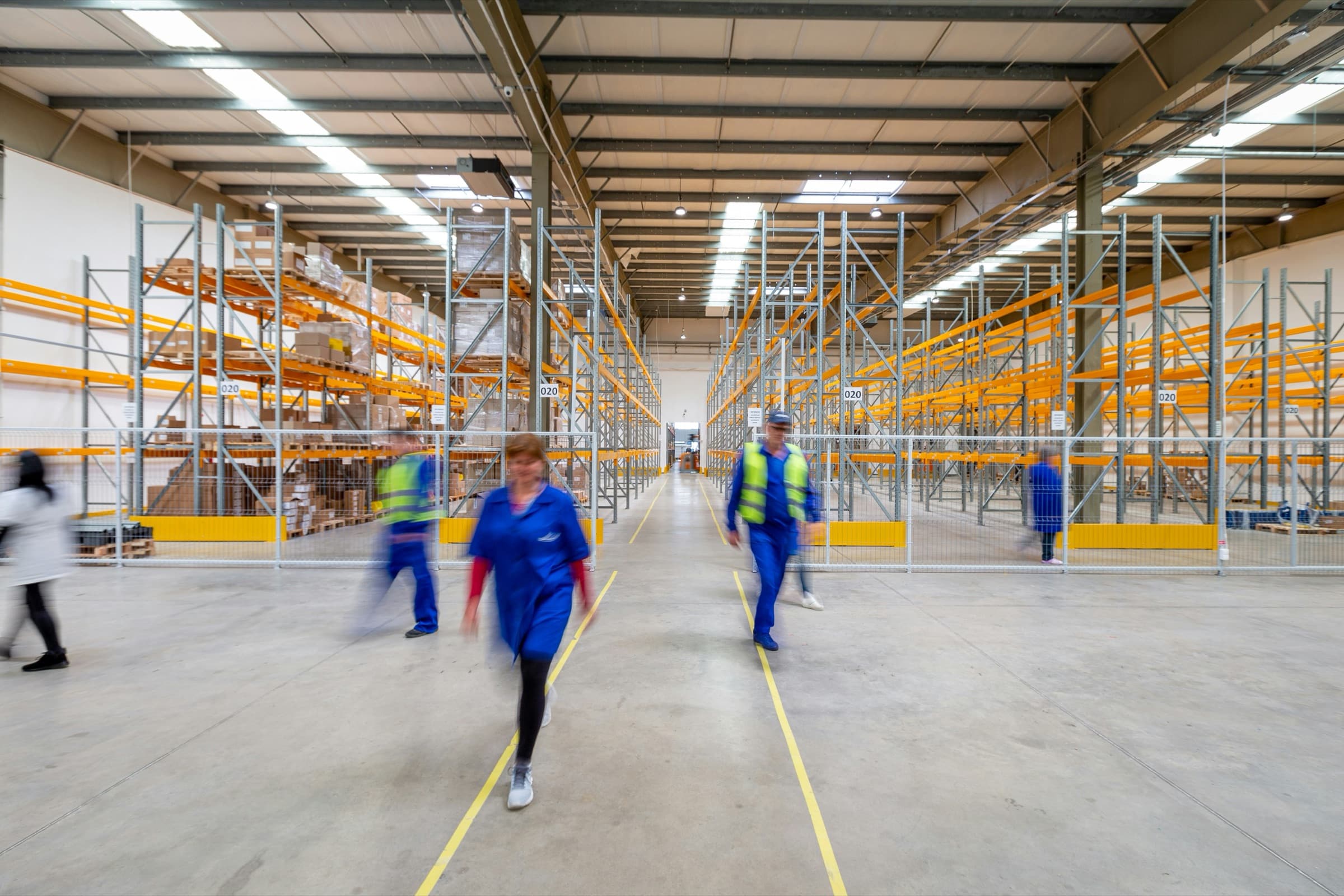 Warehouse staff in overalls and hi-vis vests crossing a busy distribution-centre floor.