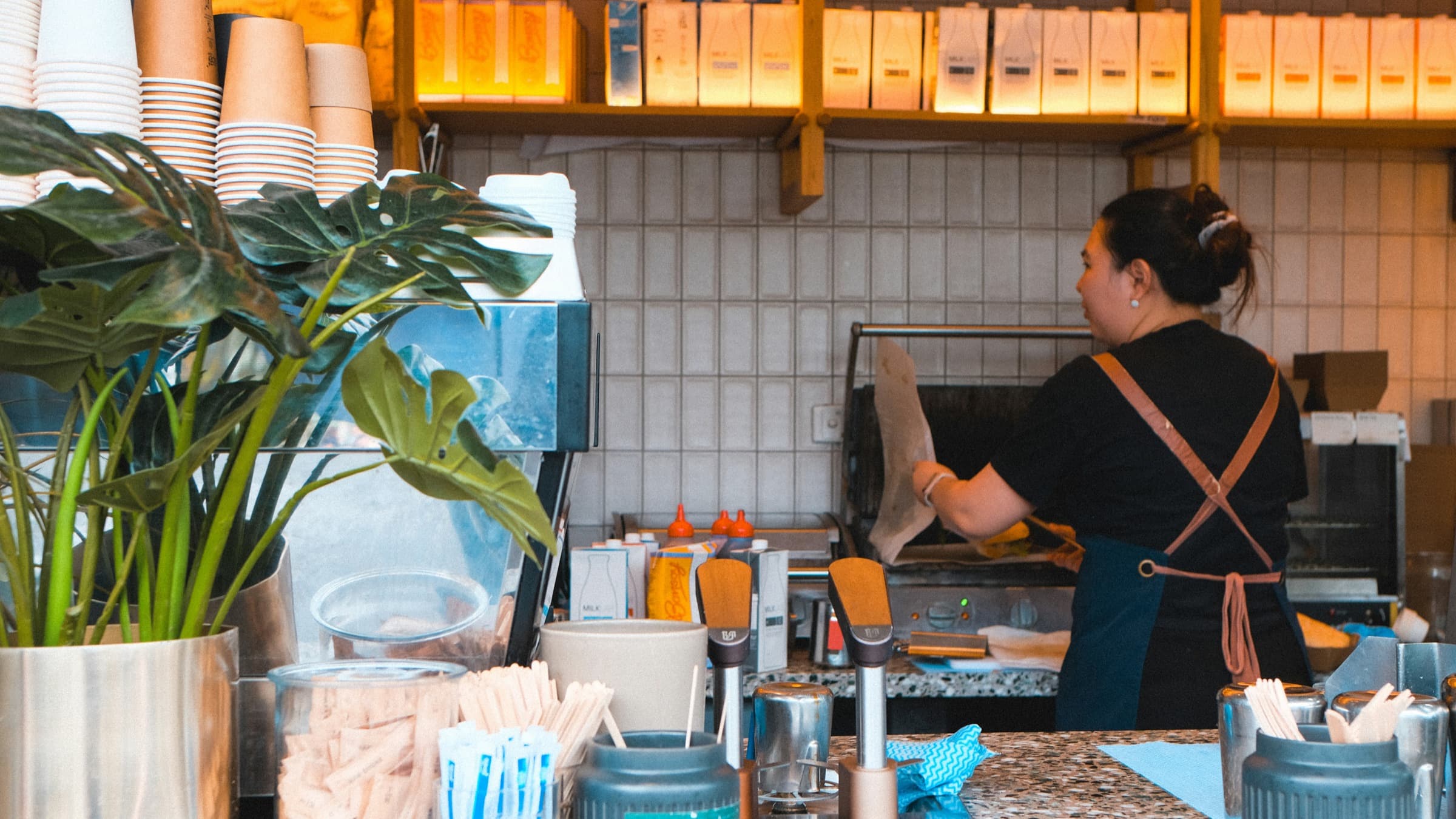 A café worker in an apron at the grill behind a counter stacked with takeaway cups.