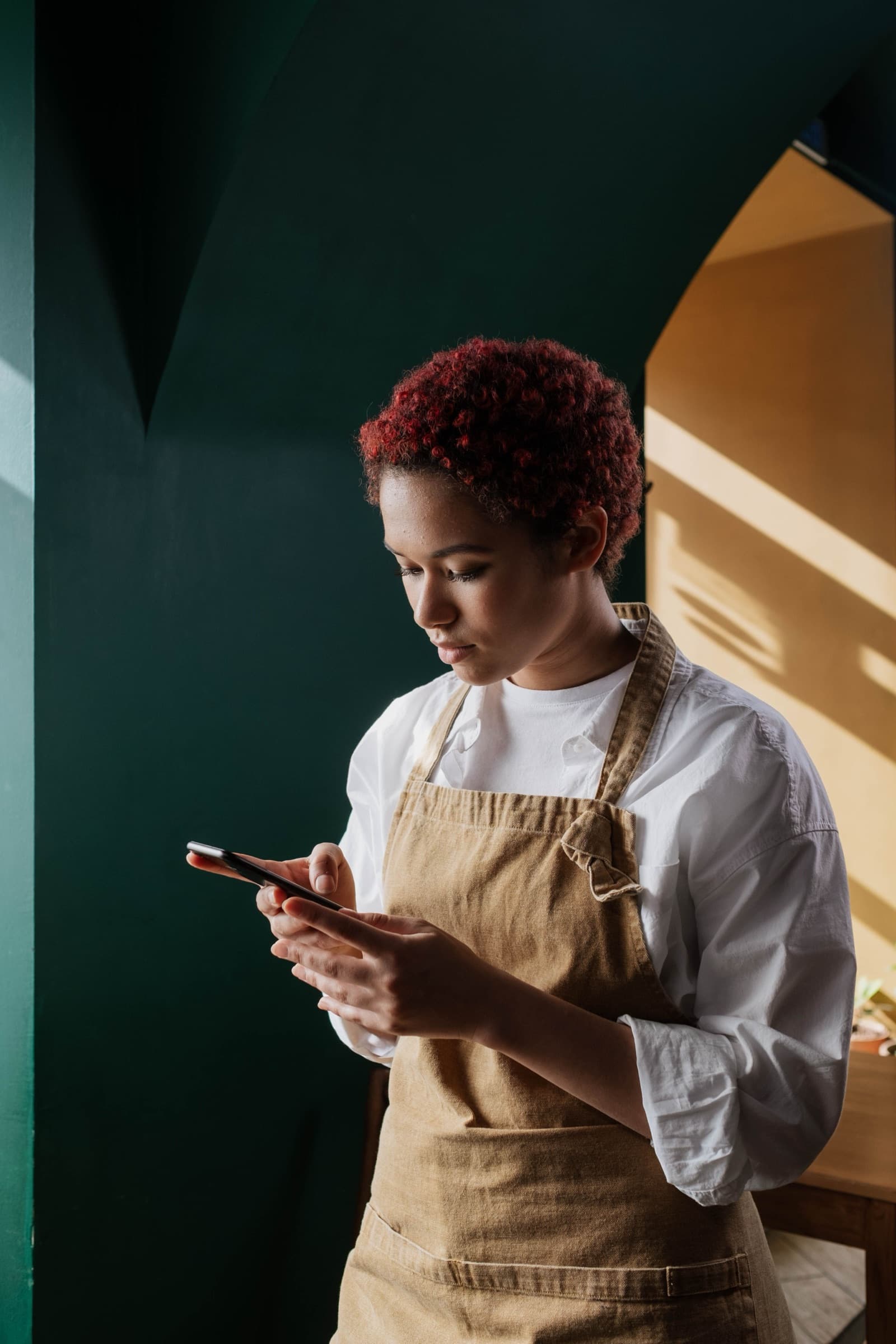A hospitality worker in an apron tapping her phone beside a green wall.